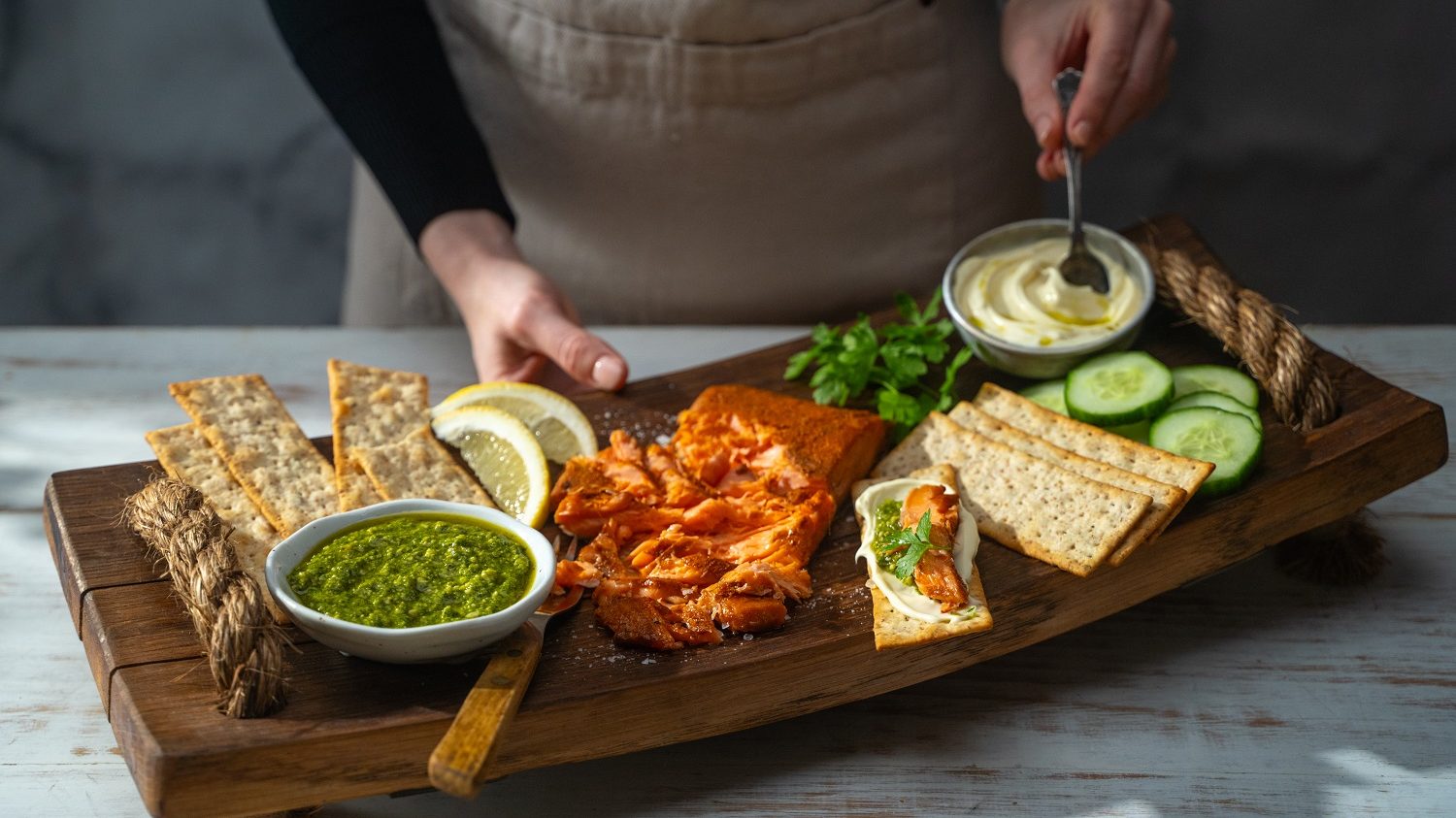 A person serving a wooden tray of salmon, crackers, pots of sauces and cucumber slices on it.