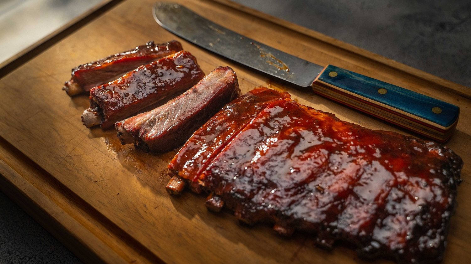 A rack of cooked and saucy pork spare-ribs on a wodden board with a large knife.