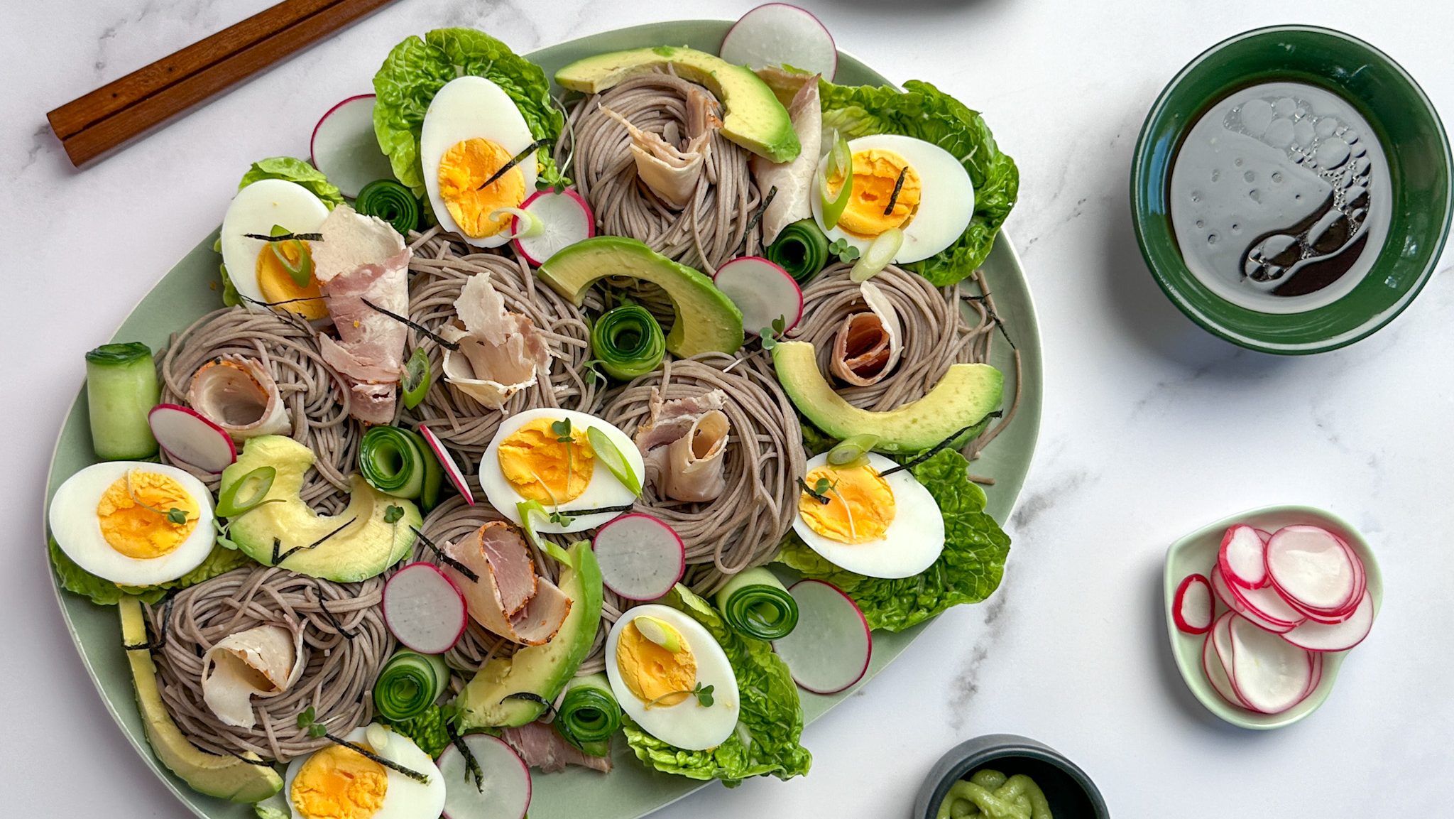 A platter of soba noodles, boiled eggs, avocado, radish and cold meat with small bowls of condiments and sauce.