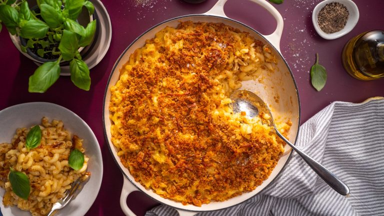 A pot of macaroni cheese with brown crumbs, a serving in a bowl and basil plant.