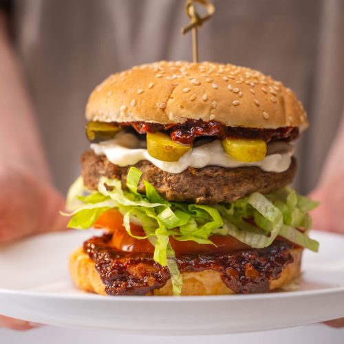 A person is offering a tall beef burger with tomato, lettuce and Gurkin on a white plate.