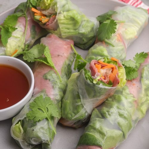 Six beef summer rolls on a plate with a bowl of red dipping sauce.