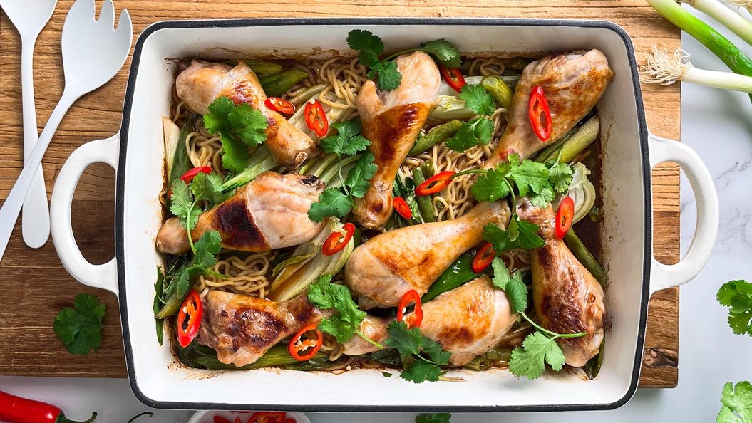 Chicken drumsticks and noodles baked in a rectangle white oven tray, garnished with red chill and green herbs.