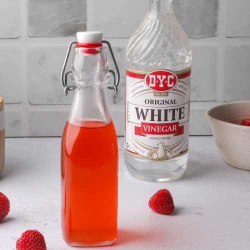 A bottle of red liquid and white vinegar surrounded by strawberries and a bowl of strawberries.
