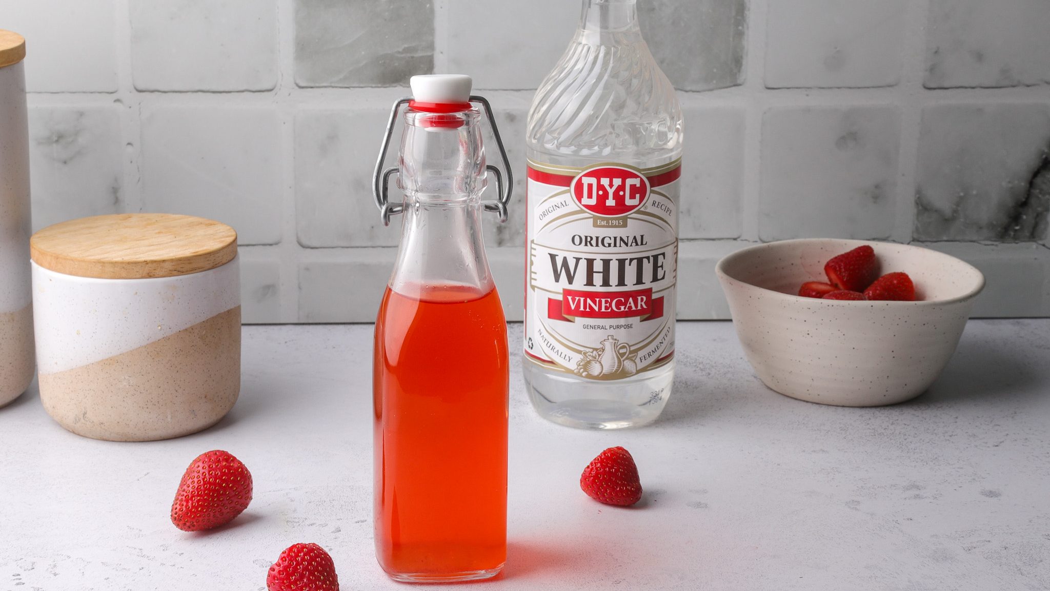 A bottle of red liquid and white vinegar surrounded by strawberries and a bowl of strawberries.