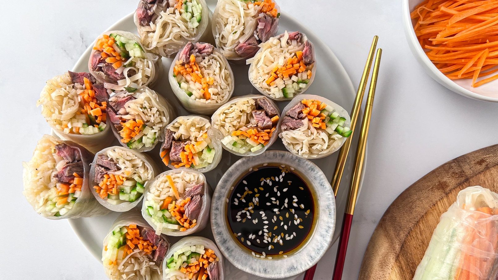 Several rice paper rolls cut side up showing colourful ingredients on a plate with a small bowl of dipping sauce and chopsticks.