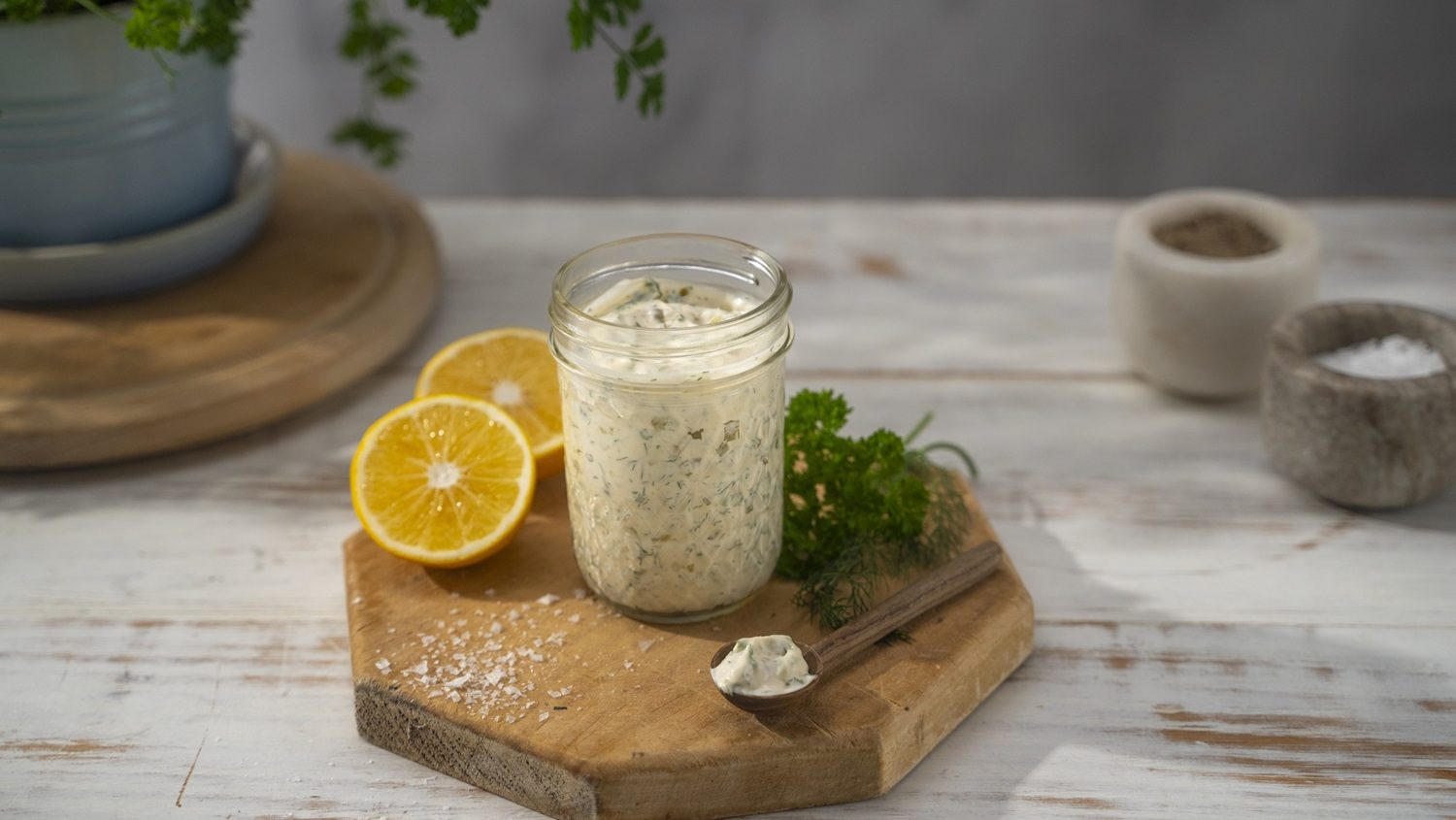 A jar full of whiteish sauce with lemon halves, parsley and garlic by its side on wooden board.
