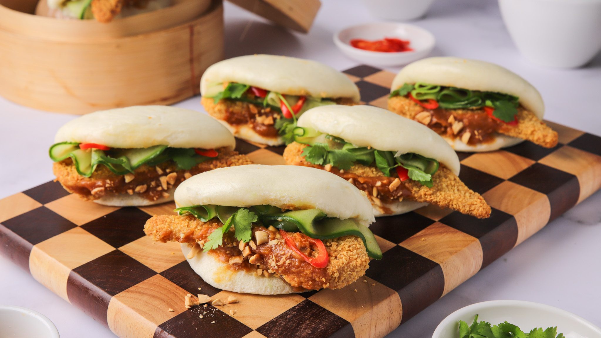 Close-up of fluffy bao buns filled with juicy, golden-brown chicken tenders drizzled with savory satay sauce, garnished with fresh herbs and a sprinkle of sesame seeds