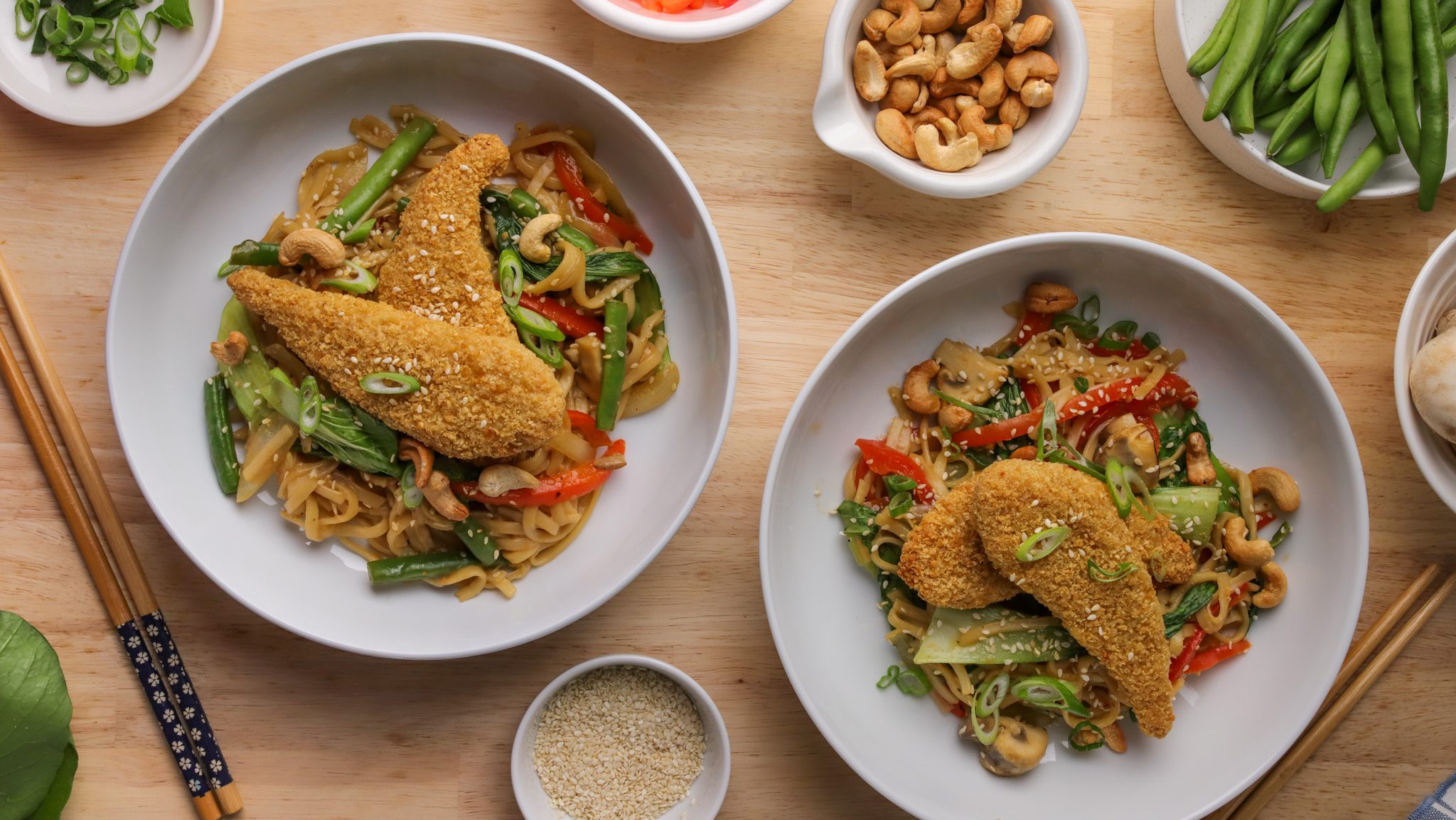 Two bowls of stir-fried noodles topped with crumbed fried food. Small bowls of condiments and a pair of chopsticks.