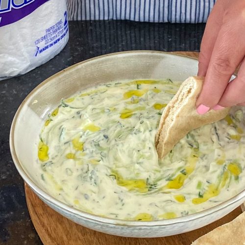 Pale green coloured creamy dip with a hand holding a piece of flat bread, a packet of kitchen towel in rear.