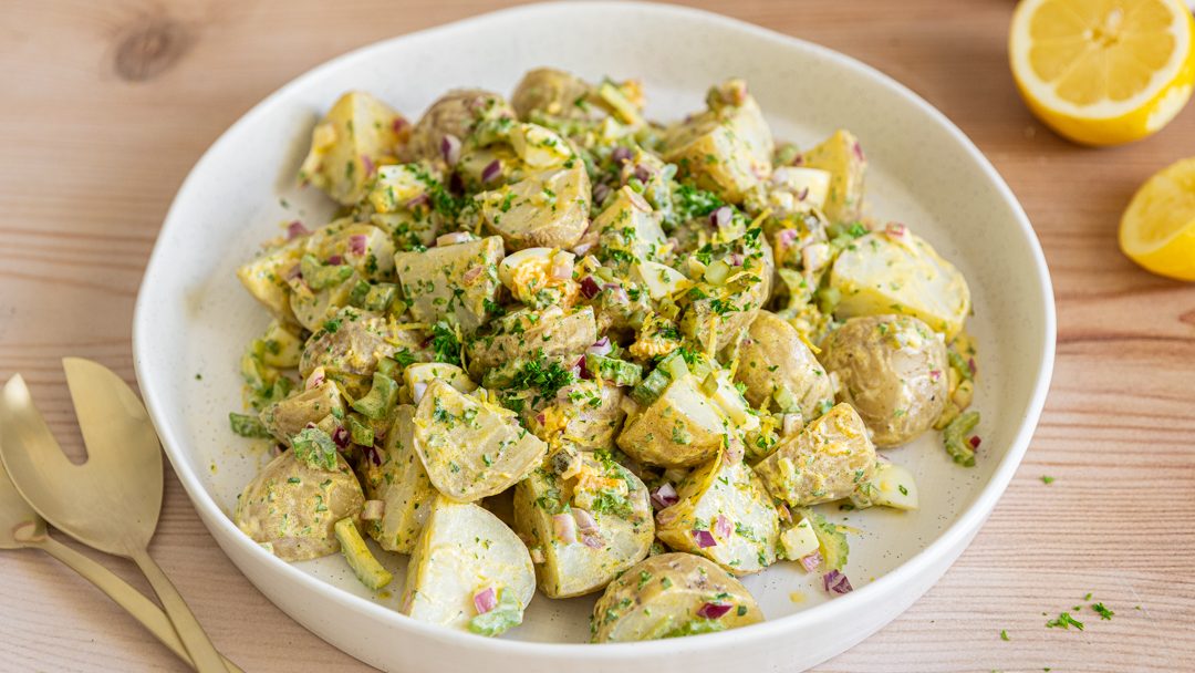 A white bowl of potato salad, lemon halves and salad servers.