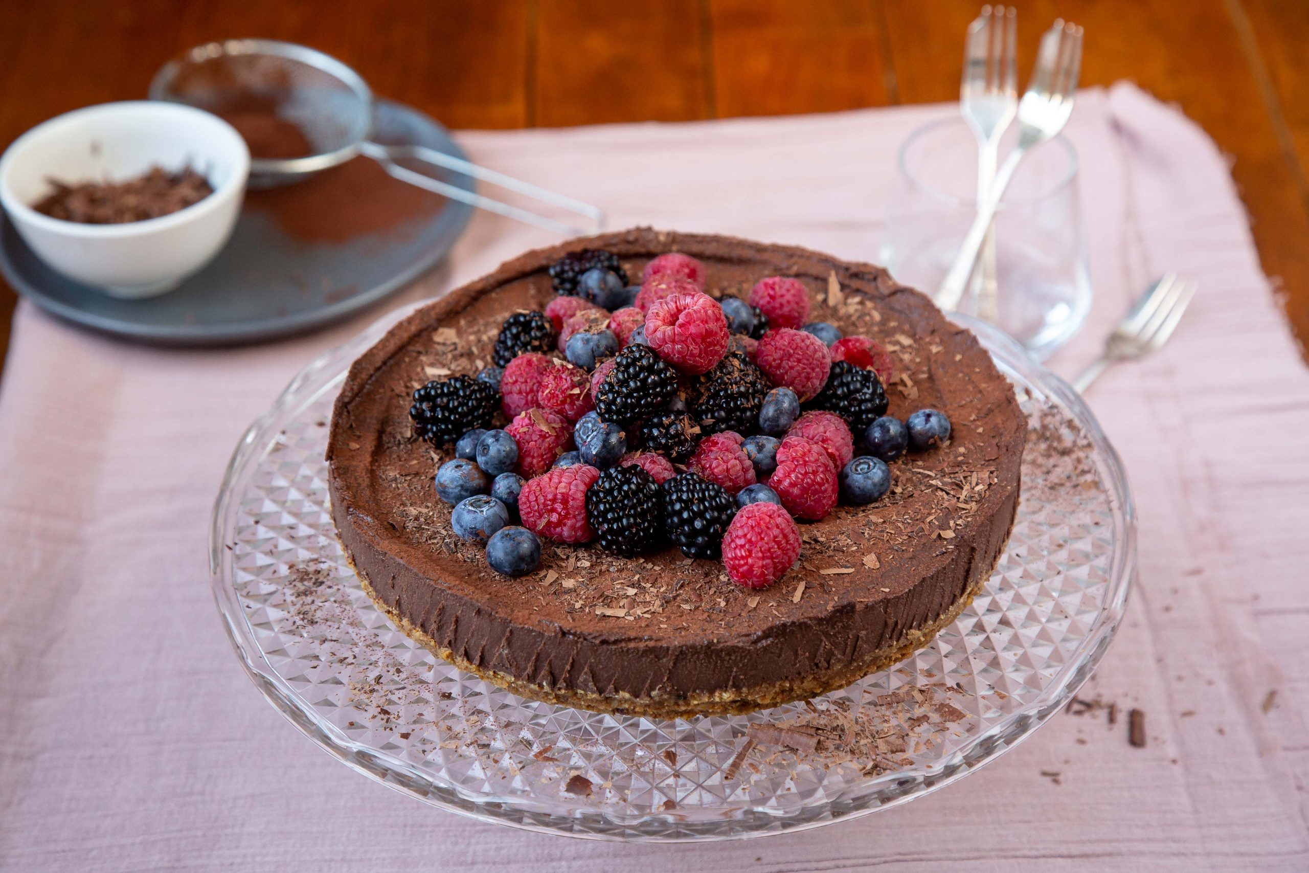 Chocolate cheesecake sits with blueberries and raspberries sprinkled on top sitting on a glass cake stand.