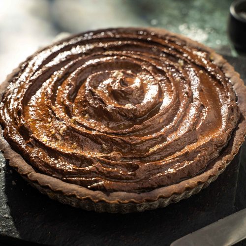 A whole round chocolate cream pie on black slate on dark green table with a knife.