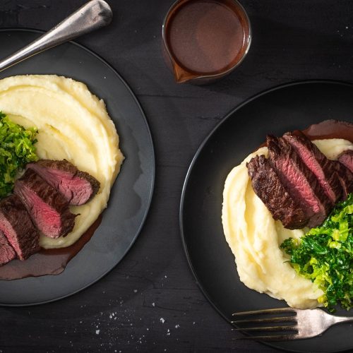 Two black plates of steak slices on white mash and greens on black table with a pot of chocolate sauce.