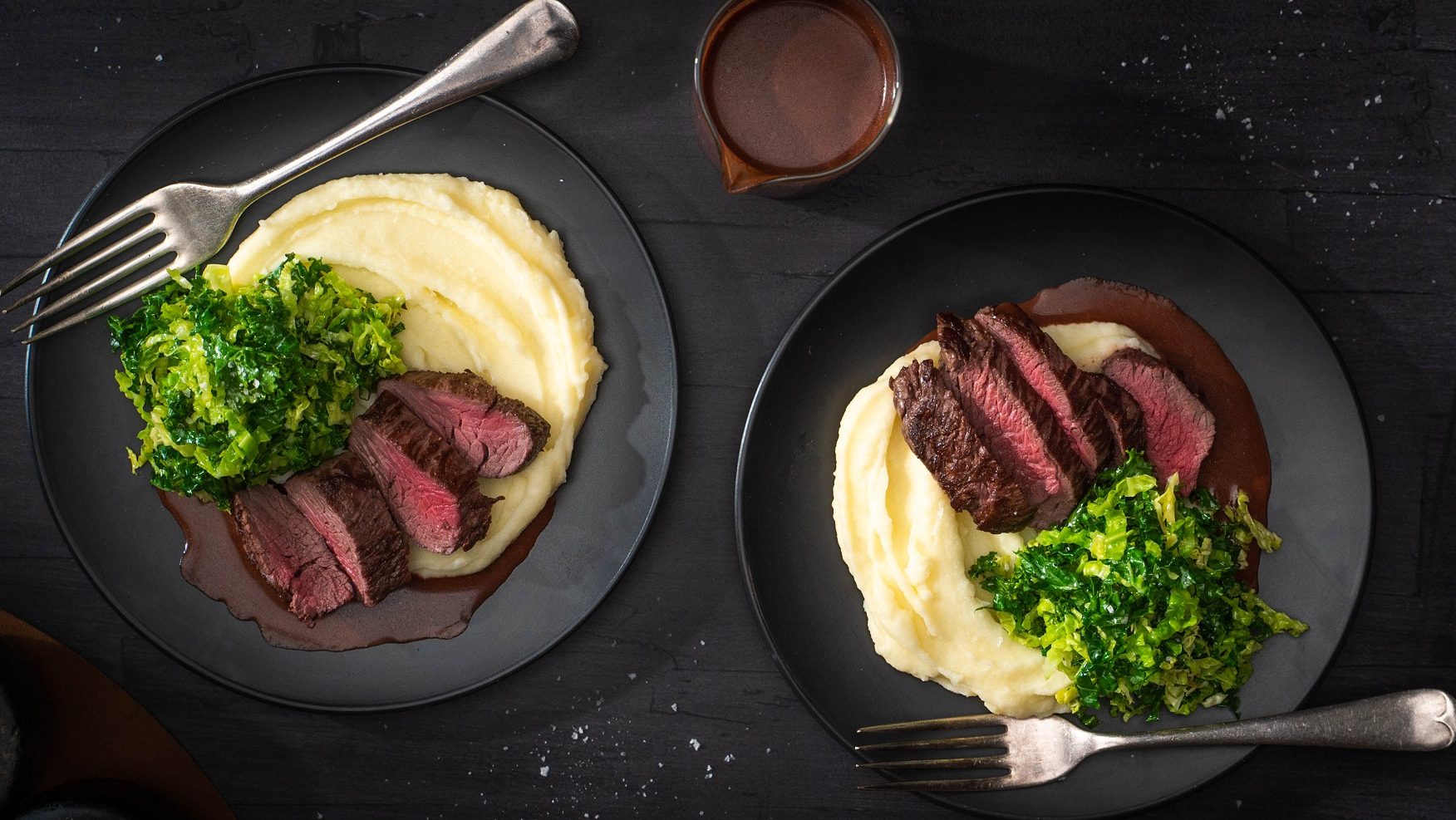 Two black plates of steak slices on white mash and greens on black table with a pot of chocolate sauce.