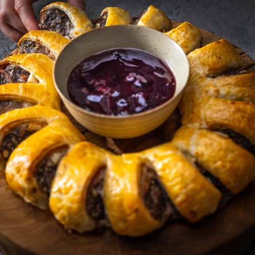 A baked sausage roll ring with a bowl of purple dipping sauce in the middle on round wooden board. A hand is grabbing one portion.