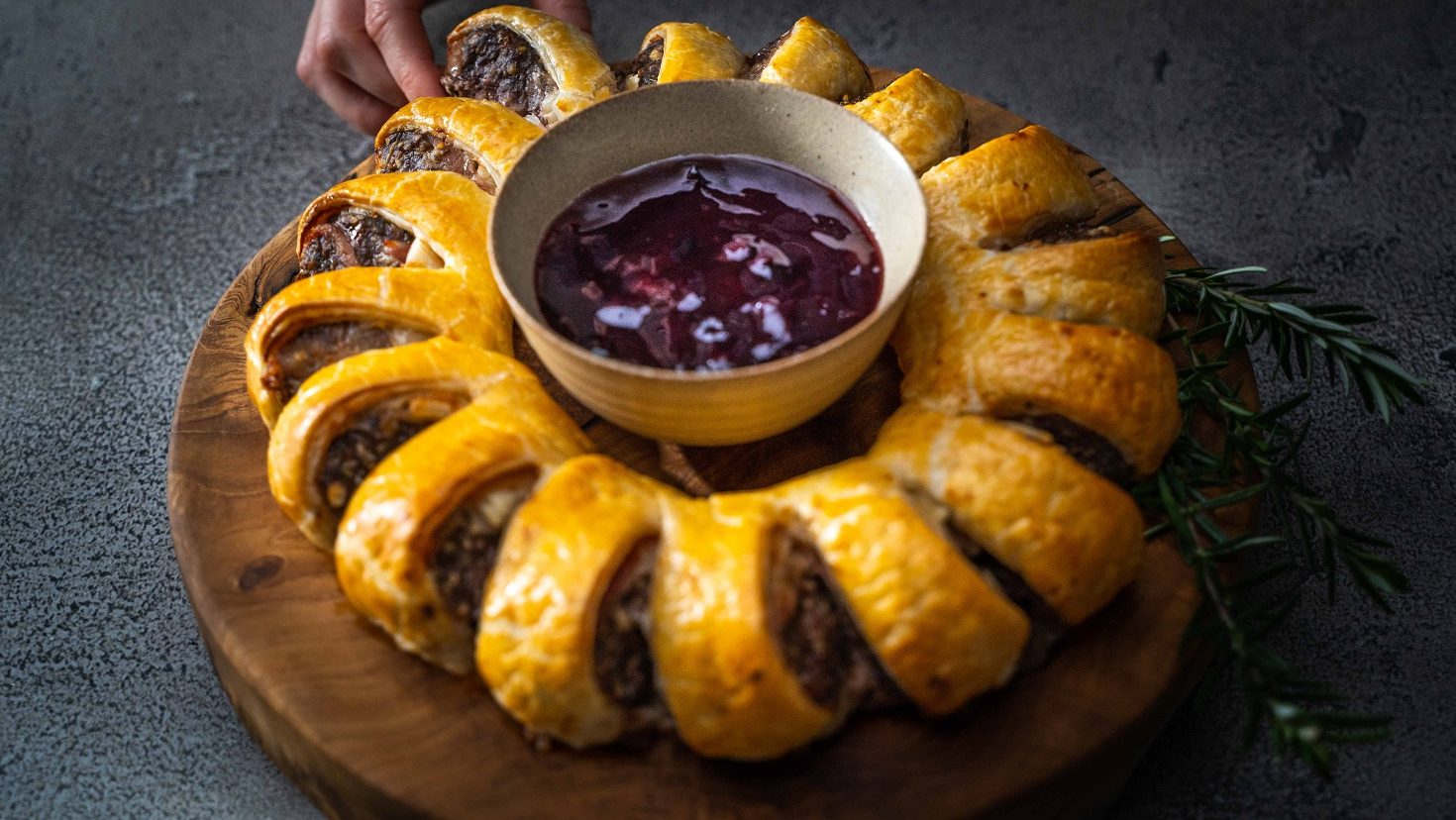 A baked sausage roll ring with a bowl of purple dipping sauce in the middle on round wooden board. A hand is grabbing one portion.