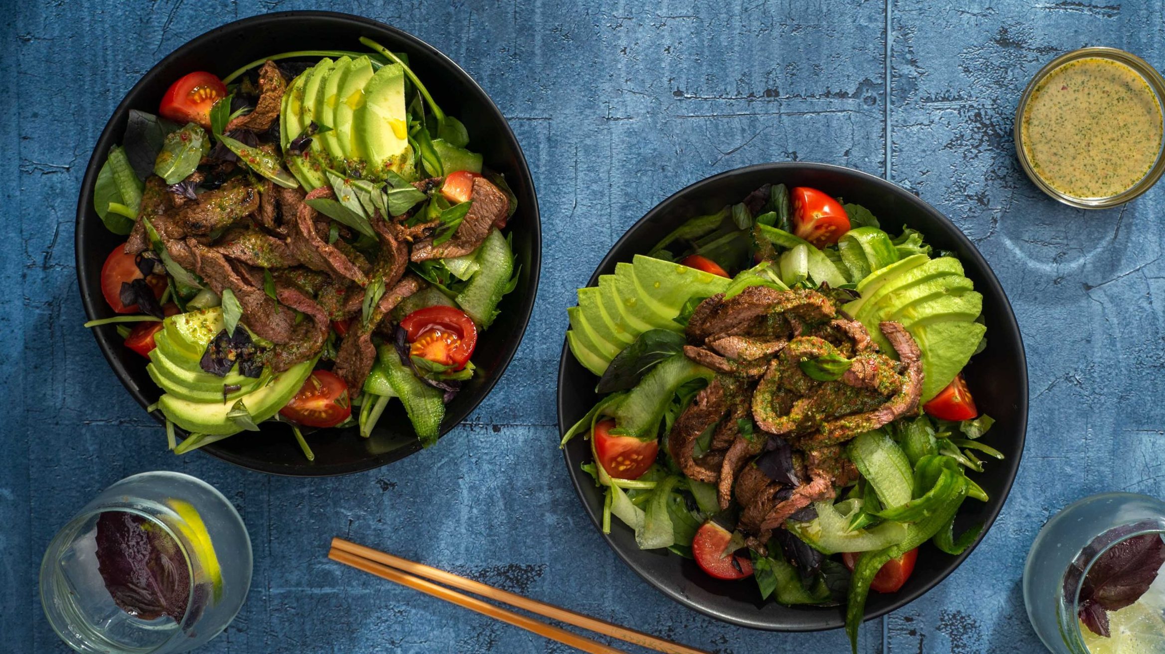 Two black bowls of salad with avocado and meat strips.