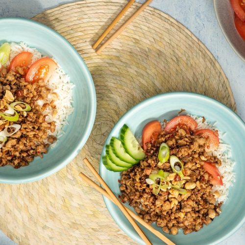 Two blue bowls of meat, tomato and cucumber on rice on a round light brown mat with chopsticks. Tomatoes and nuts around them.