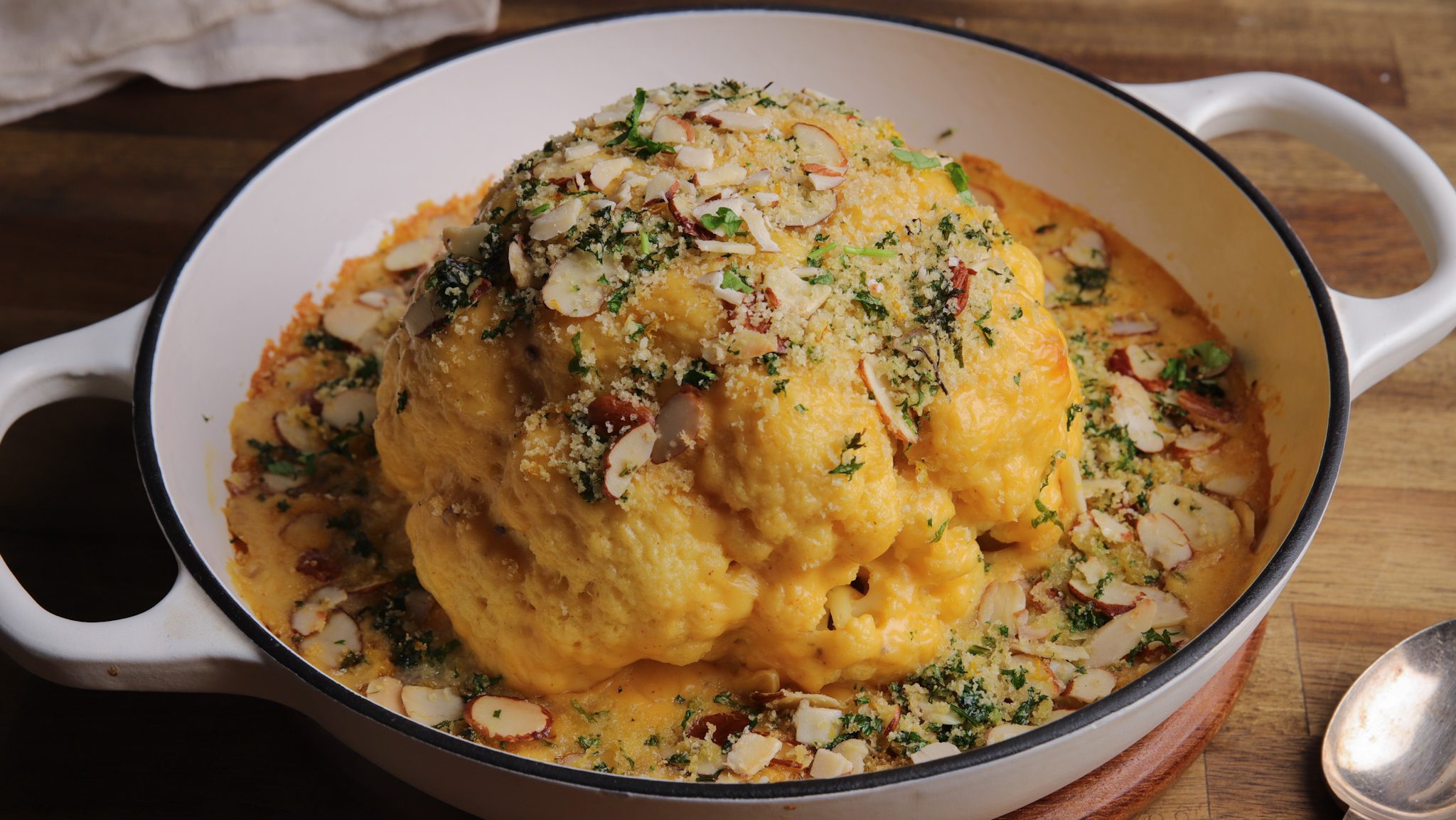 A whole cauliflower in a white pan with yellow sauce, almond flakes, parsley and crumbs on top.