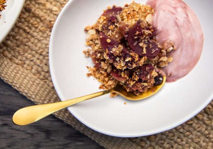 A bowl of fruit crumble with pink yoghurt in white bowl with gold spoon