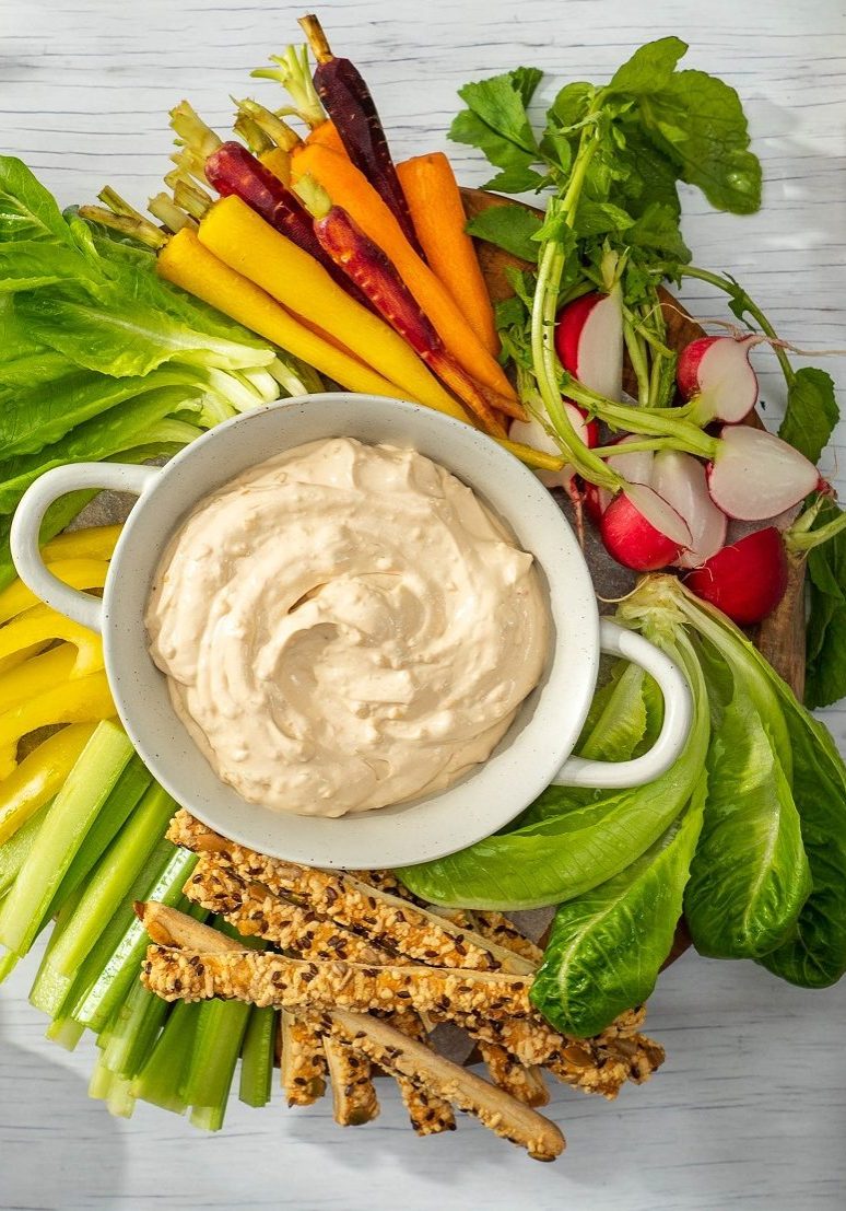 Bowl of creamy dip in the centre of a platter with colourful raw vegetables, a bowl of potato chips.