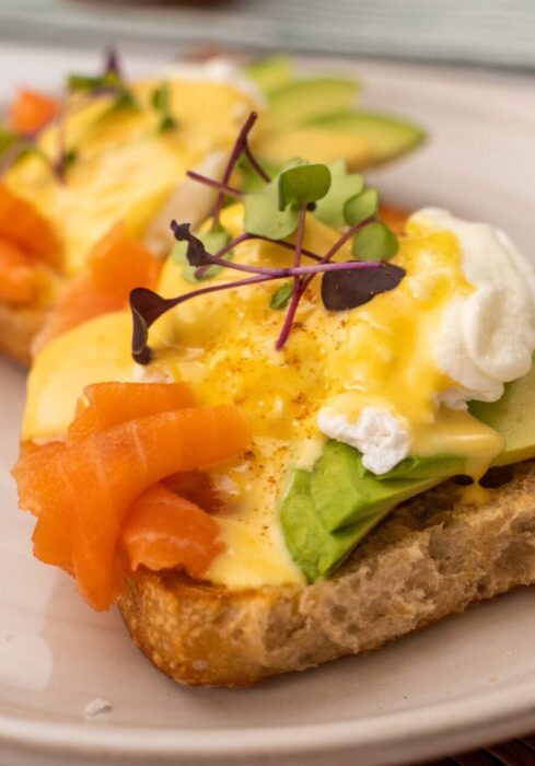 Egg with yellow sauce and herb on top of avocado slices, salmon on bread base on a white plate