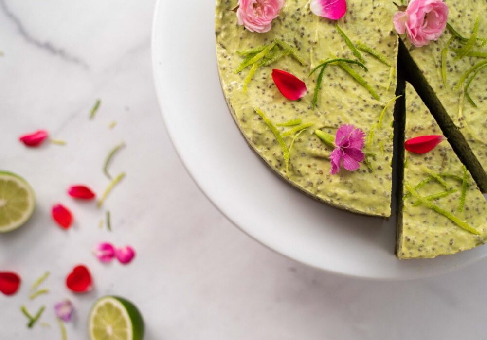 Round green cake with a slice cut topped with flowers on marble, lime halves and more flowers scattered.