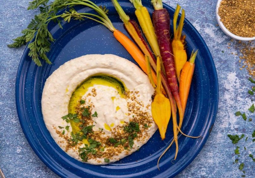 Hummus paste and carrots on a blue round plate on blue surface with a pot of brown dukka.