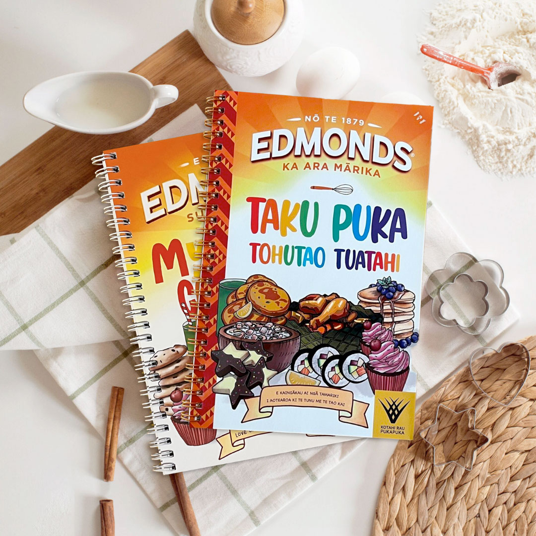 Copies of Edmonds My First Cookbook in both English and te re Maori on against a background with forks and herbs