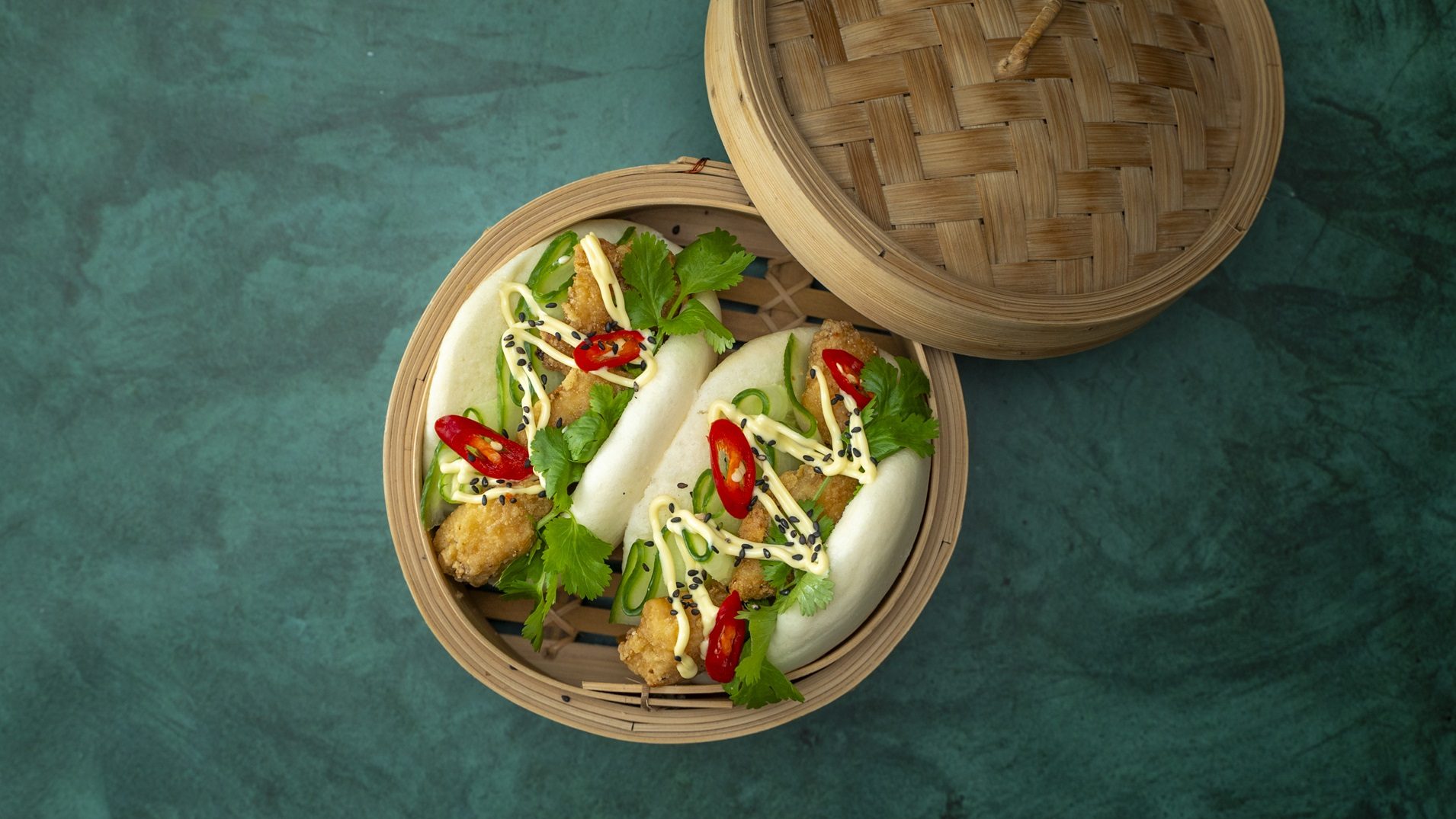Two filled bao buns inside bamboo steam basket with the lid next to it.